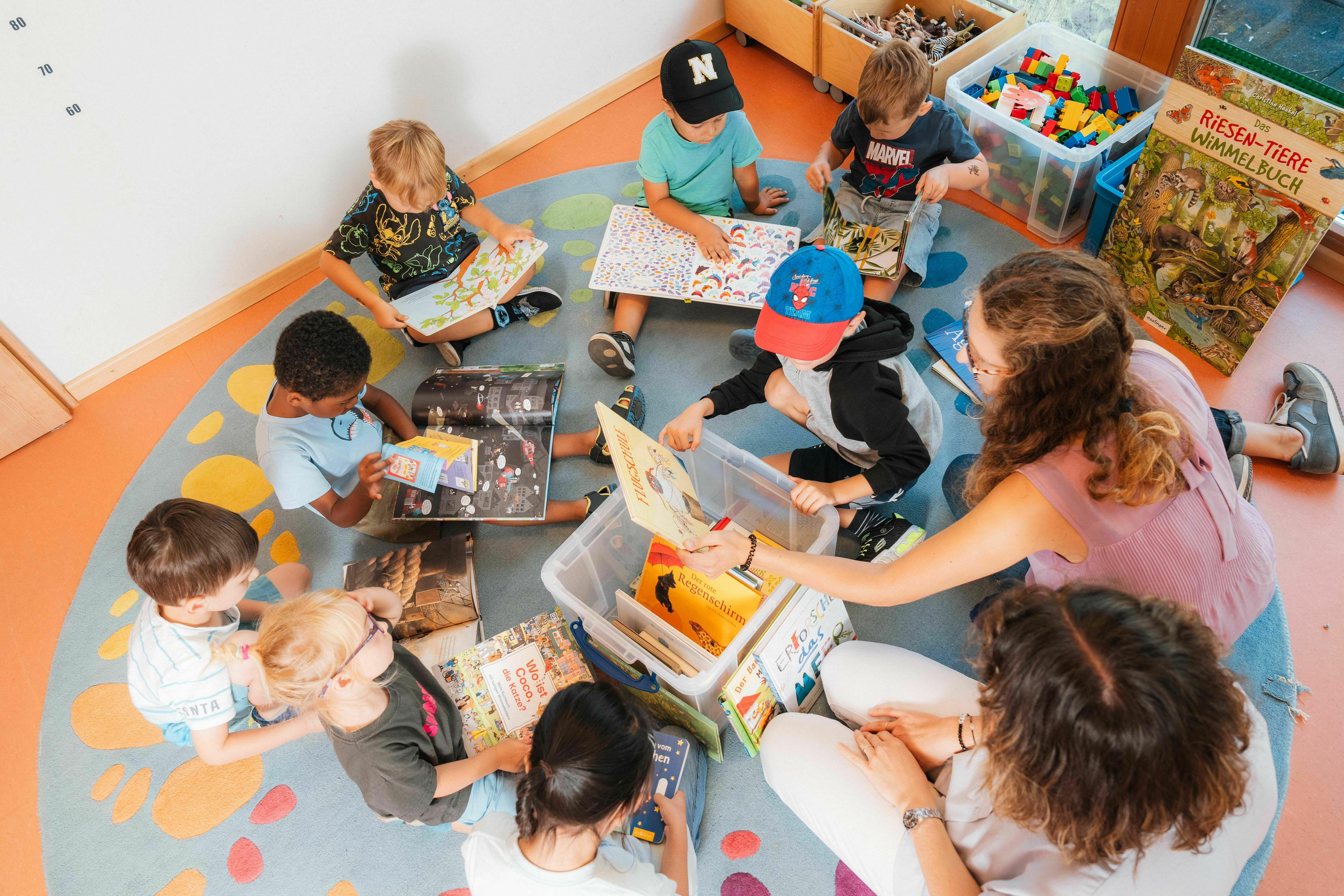 Mit verschiedenen Lese- und Lernmaterialien sitzen Kita-Kinder gemeinsam auf einem Teppich. Eine pädagogische Fachkraft verteilt die Medien.